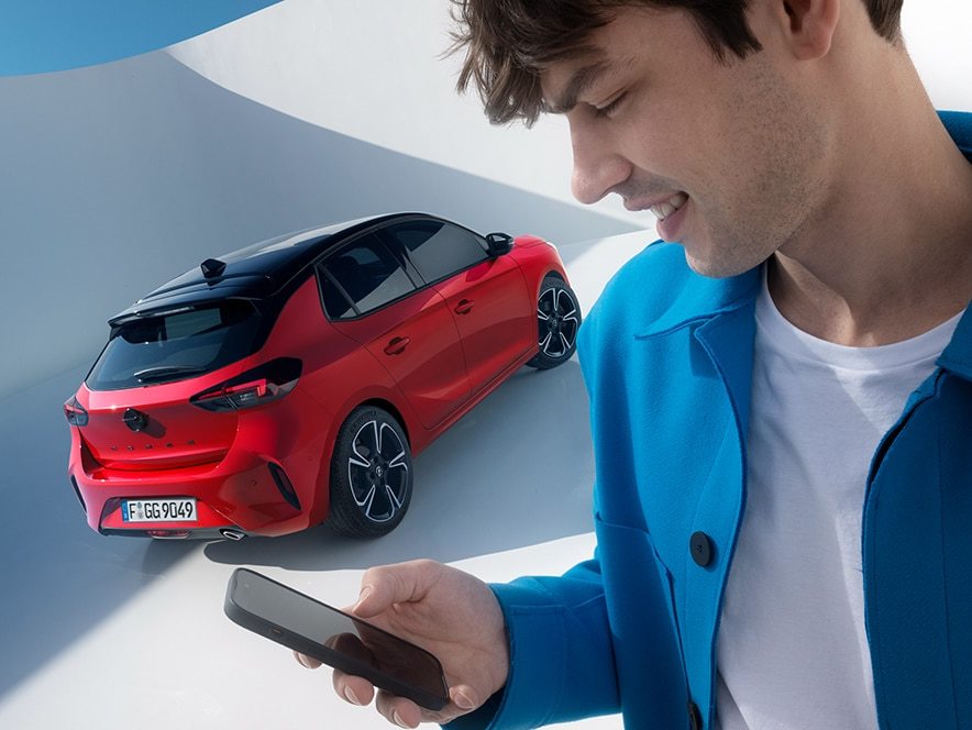Un uomo con lo smartphone in mano mentre alle spalle compare una Nuova Corsa rossa con i cerchi in lega grigio-neri